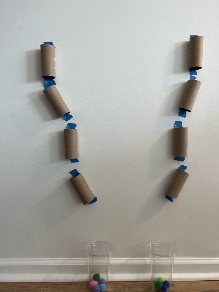 2 sets of 4 Brown toilet paper rolls taped to the wall with blue tape. Each set of the rolls are taped to the wall in a way that a small ball can be dropped into one to another down the wall into a cup at the ground.