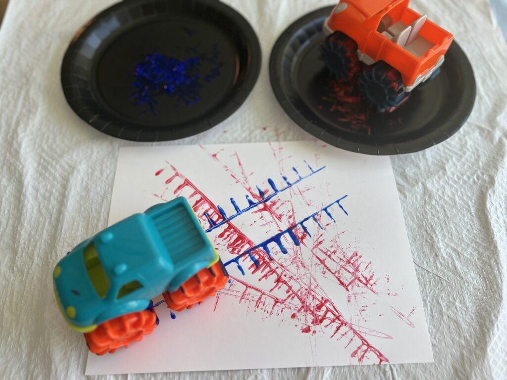 Blue paint on a black plate and red paint on another black plate. A piece of paper set in the middle. Trucks whose wheels have been cover in the red and blue paint being driven on the paper. The trucks being moved across the paper have made blue and red tire marks across the page.