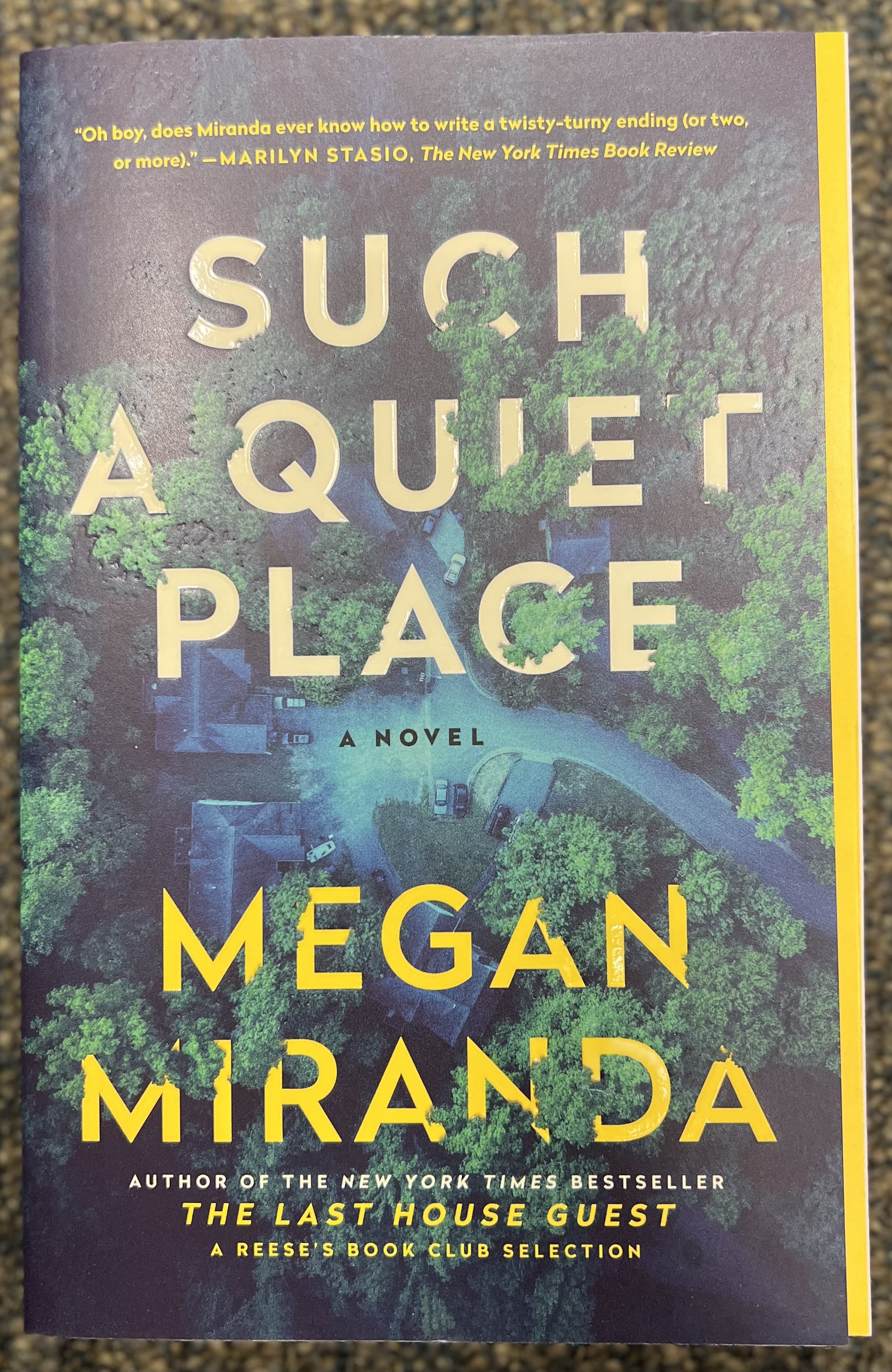 This is a photograph of the book Such A Quite Place by Megan Miranda; sitting on the ground of blue carpet.