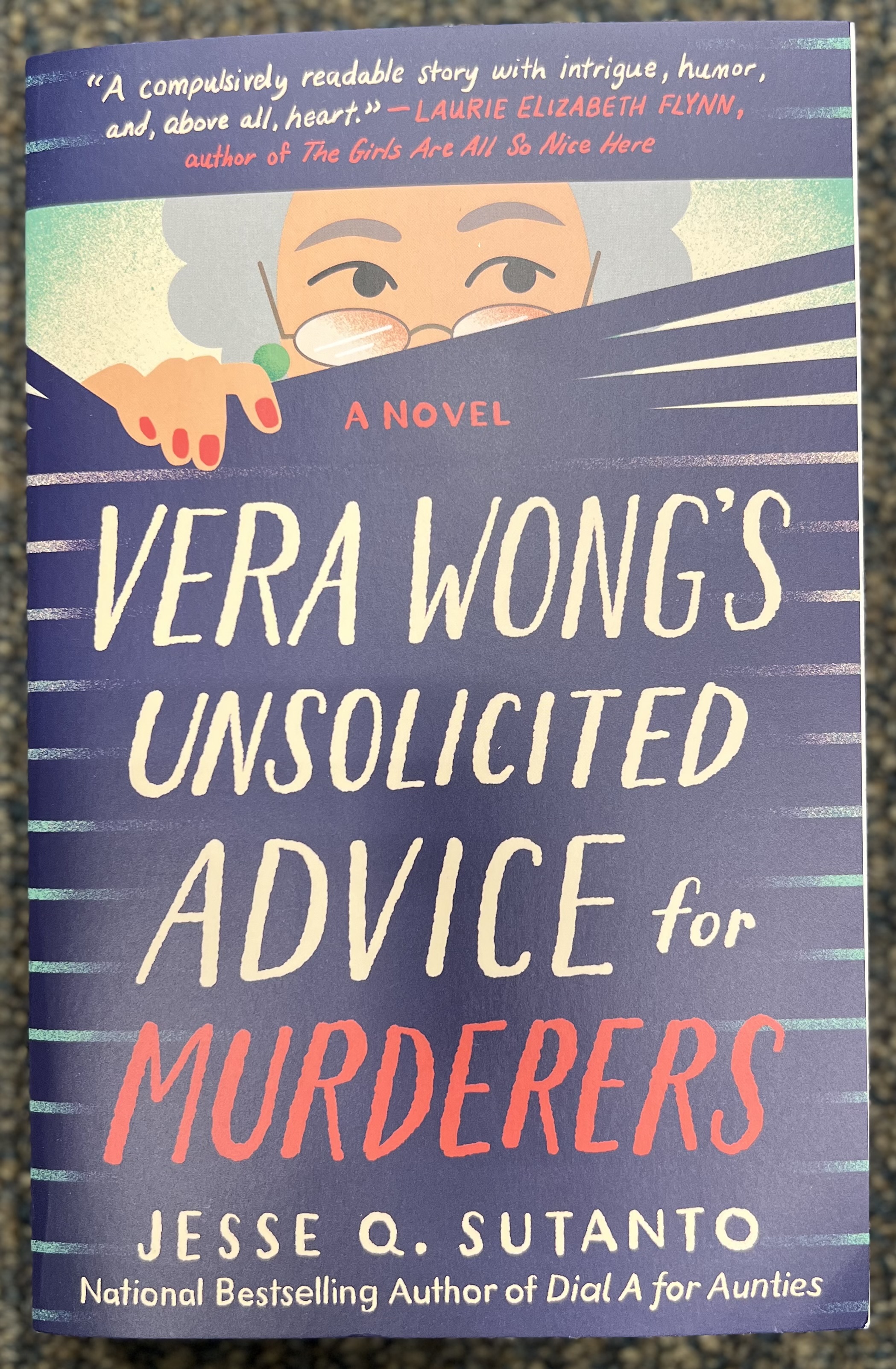 This is a photograph of the book Vera Wong's Unsolicited Advice for Murderers by Jesse Q. Sutanto; sitting on the ground of blue carpet.