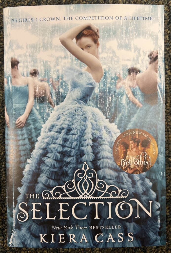 This is a photograph of the book The Selection by Kiera Cass; sitting on the ground of blue carpet.