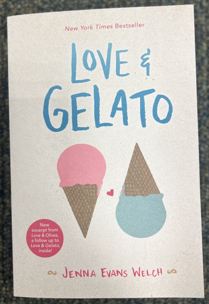 This is a photograph of the book Love and Gelato by Jenna Evans Welch; sitting on the ground of blue carpet. The book itself is a cream color with the words "New York Times Bestseller" in pink and small print. Below that in big blue print are the words " Love & Gelato". Below that are two ice cream cones right next to each other in the same size with a small pink heart in between them. The ice cream cone in the left has pink ice cream and a brown cone. The The ice cream cone in the right has blue ice cream and a brown cone. At the very bottom of the cover are the words "JENNA EVANS WELCH" in pink font.  