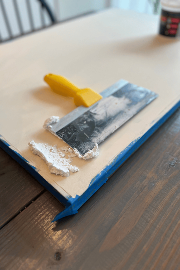 A canvas painted a cream color sitting on a gray table with blue painter's tape. On the canvas is a lump of spackling with a spackling tool with a yellow handle on top on top of the spackling.