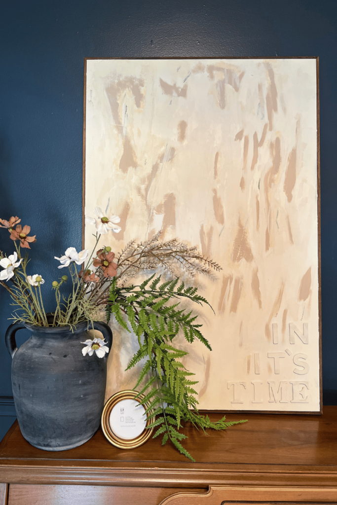 The after picture of the repurposed canvas. The canvas is leaning against a dark blue wall sitting on brown wood piano. The canvas has a brown wood frame. The canvas is painted a cream color with abstract darker cream color with the words "In its time" in the right corner. Along with the canvas on the piano it is paired with a black vase with flowers and greenery coming out of it and a small gold frame with no picture in it.