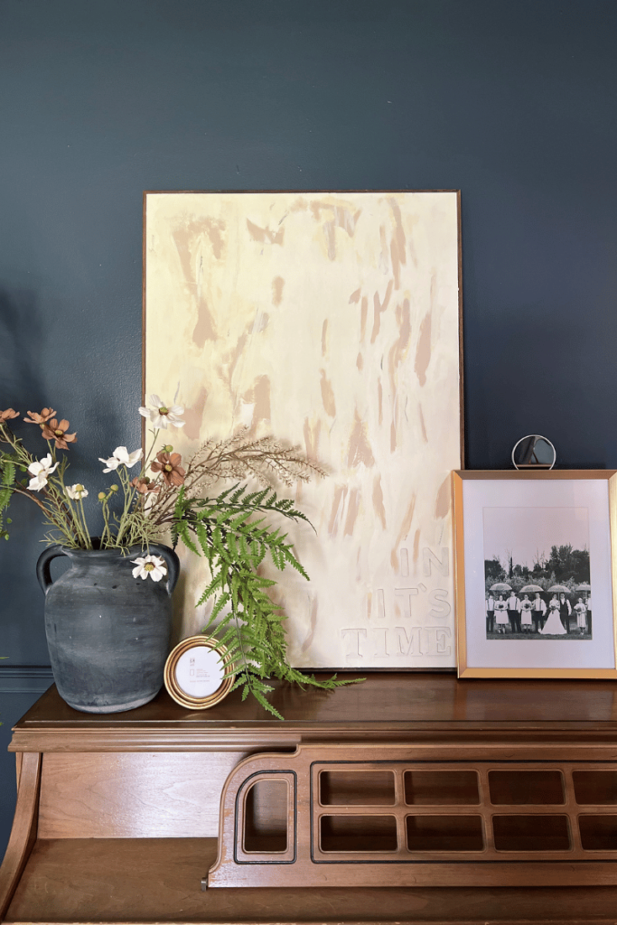 Picture of the repurposed canvas. The canvas is leaning against a dark blue wall sitting on brown wood piano. The canvas has a brown wood frame. The canvas is painted a cream color with abstract darker cream color with the words "In its time" in the right corner. Along with the canvas on the piano it is paired with a black vase with flowers and greenery coming out of it and a small gold frame with no picture in it to the left of the canvas. On the right of the canvas is 8 by 10 gold frame with a wedding picture in it.