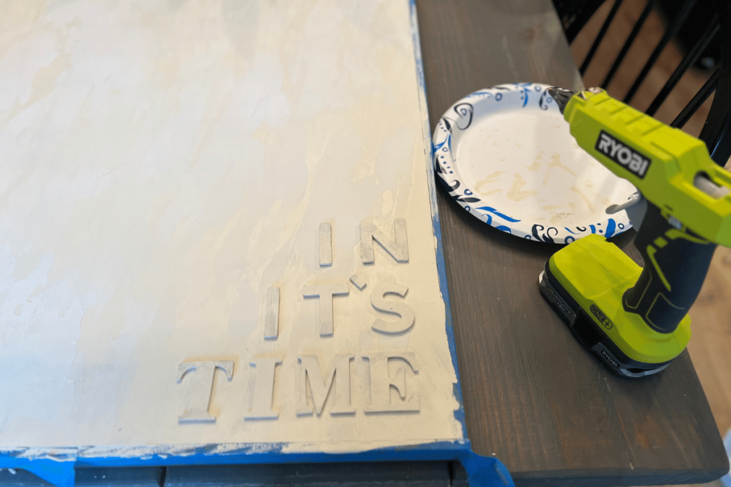 A picture of the bottom right corner of the canvas with blue painter's tape on the sides. In the corner a glued on words "IN IT'S TIME". The right of the canvas on the table is a lime green Ryobi glue gun and in front of the glue gun is a white plate with blue details.