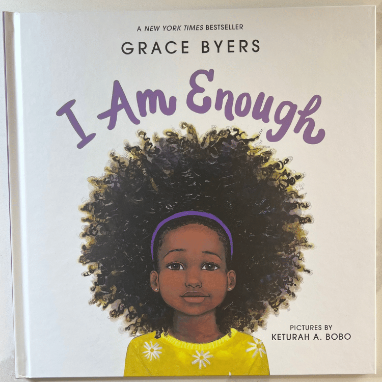 The book cover of I Am Enough sitting on a white countertop. The cover has a white background the name of the book at the top. A close up drawing of a little girl who is black with big curly hair with a purple headband and wearing a yellow shirt with white daisies  is centered in the middle of the book.