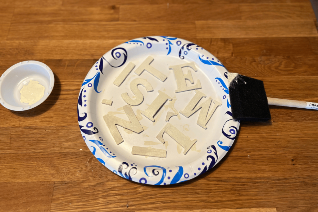 A white paper plate with blue details on the rim a butcher block counter top. On the plate are the Letters "I", "T", "E", "S", "I", "M", "T", "I", "N", and a coma. The letters and coma are painted a cream color. Leaning on the rim and laying on the counter top is a back sponge paint brush on the right. On the left of the plate is a small paint cup that is white and has cream paint inside. 