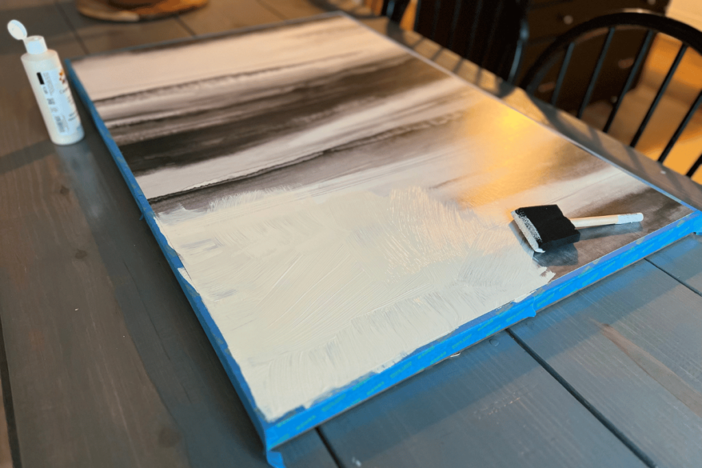 The canvas is sitting on a gray table with the wood frame covered in blue painter's tape. The canvas is black and white abstract piece and is being painted over by cream pant. Laying on the canvas is a black sponge paint brush sitting next to what has been painted. 