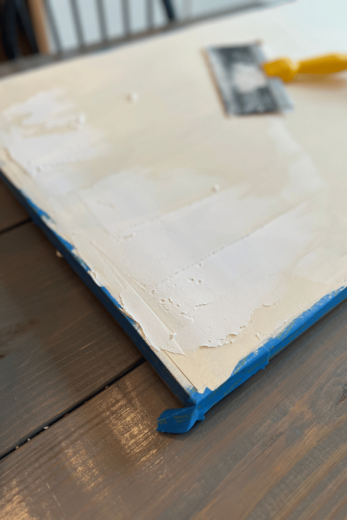 A canvas painted a cream color sitting on a gray table with blue painter's tape. On the canvas is spackling smeared on the top side with a spackling tool with a yellow handle off to side bleared to the right on the canvas.