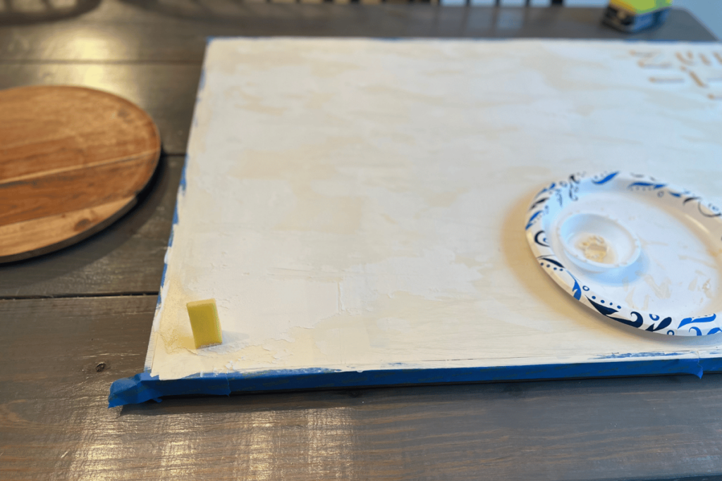 Picture of the painting the repurposed canvas on a gray table. A paper plate that is white with blue detail on the rim is sitting to the right on the canvas. On the plate is a small paint cup with cream paint in it. On the left bottom corner is a yellow sponge starting to paint the canvas. The canvas has a blue painter's tape covering the frame. The canvas is painted a cream color with the words "In its time" in the right corner.