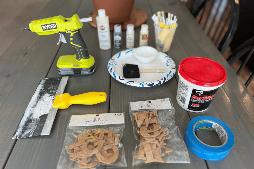 A picture of all the supplies used to repurpose the canvas. In the picture all the items are setting on the table. There is plastic cup full of paint brushes and sponges, 3 small tubes of paint and 1 large tube of paint in cream. A lime green Ryobi battery powered glue gun, a paper plate with blue detailing with a sponge paint brush and small paint cups on it. A tub of spackling with a red top. A spackling tool with a yellow handle. Two packages with lettering made out of cork. A Roll of blue painters tape.