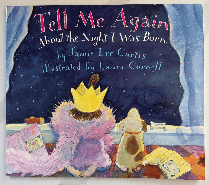 A photograph of the book cover of Tell Me Again About The Night I Was Born sitting on a white counter top. The cover of the book is the back of little girl with brown hair with a yellow crown on her head wearing pink boa and a pink dress with a tan dog with brown spots sitting right next to her. The little girl and dog are sitting in front of a large window looking out into the stary night sky. The words "Tell Me Again" in pink "About the Night I Was Born" in white centered at the top of the cover and the words "by Jamie Lee Curtis illustrated by Laura Cornell" in blue right under the other wording.