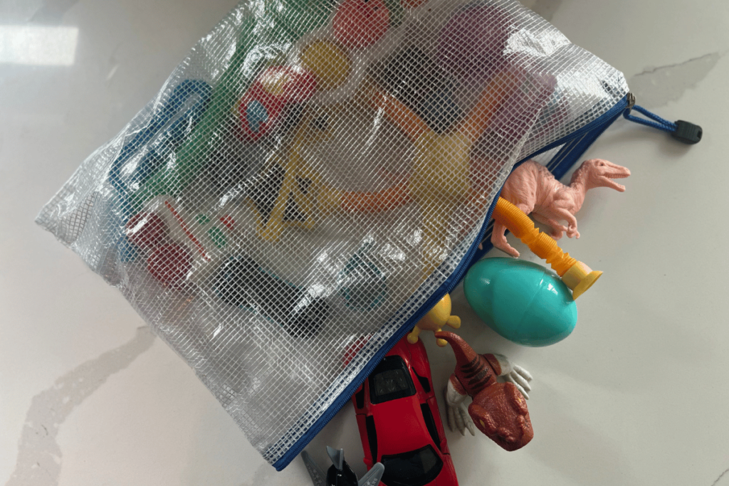 A mesh zipper bag with blue zipper detailing and the bag unzipped. Spilling out of bag are a bunch of little small toys. A peach dinosaur, blue easter egg, brown and white dinosaur, and a red car you can see spilling out on the counter top it is sitting on.