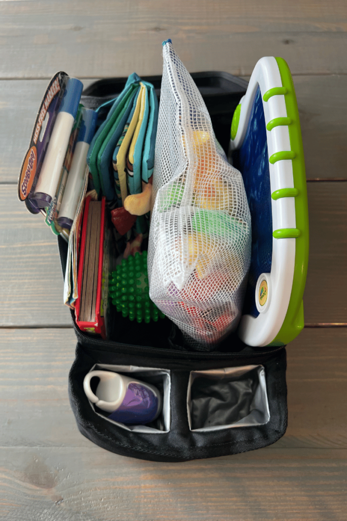 A black bin that can attach to car's seat with a seat belt with picture taken on a table. The bin is filled with two invisible pin coloring books, a green ball, a red book, two small books, a fabric book, a green and blue light up finger doodle board, a large bag of toys. With two cup holders lined in gray.