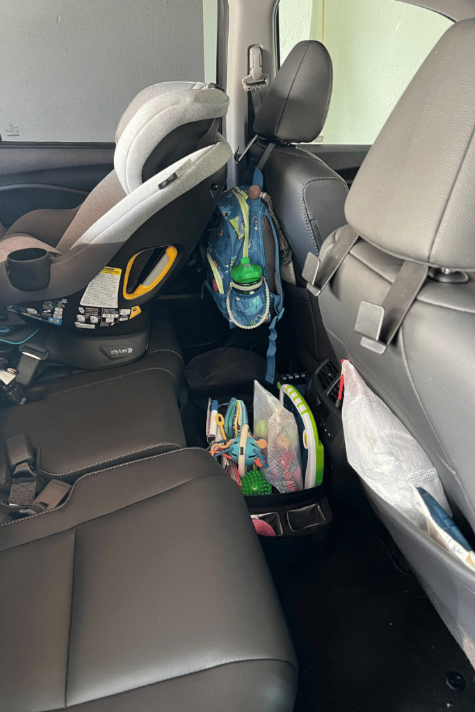 Picture of the back row of my car. With a car seat installed in the seat behind the diver's seat. A mesh zipper bag and a bag of wipes in the pocket of the back pocket of the passenger seat. Two bag hooks on the back of the diver's seat and the passenger seat with a diaper bag hanging from a hook on the diver's seat. A black bin full of different activities for a toddler is seating in the middle of the floor in-between the diver and passenger.