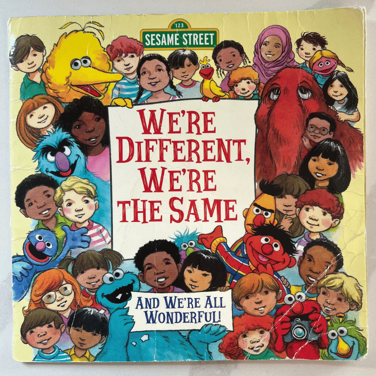 Photograph the cover of the book "We're Different, We're The Same" sitting on a white countertop. The background is yellow with drawings of big bird, Bert, Ernie, Elmo, and lots of different children all surrounding the a piece of paper with the title of the books and Cookie Monster holding a piece of paper that has written "AND WE'RE ALL WONDERFUL".